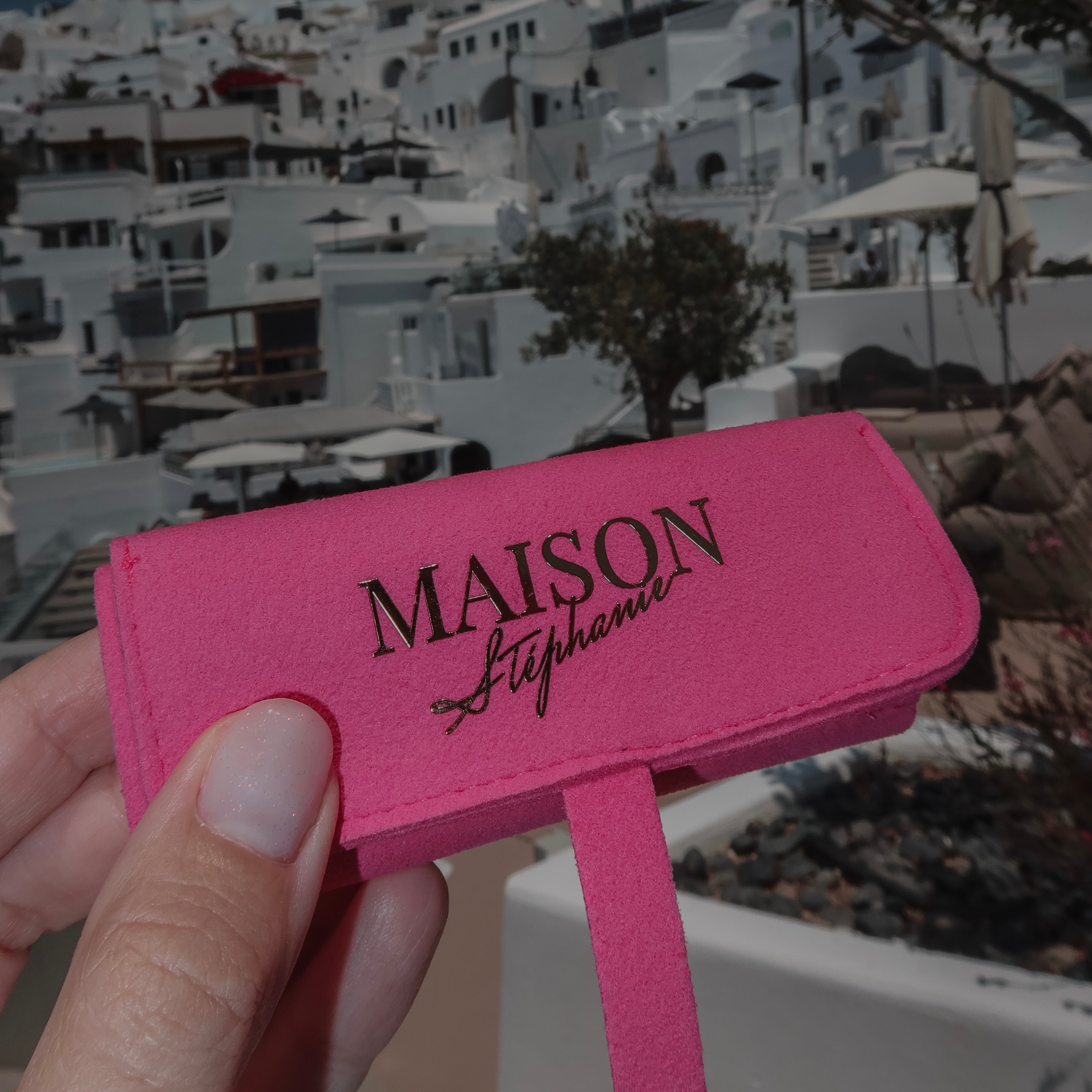 Organizator pentru bijuterii MAISON Stéphanie Travel Pouch Fuchsia, fotografiat în Oia, Santorini, cu arhitectura albă specifică insulei în fundal.
